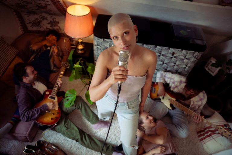 Zangeres Bieke van bovenaf gefotografeerd in een huiselijke omgeving, met in de achtergrond vier vrolijke muzikanten op giraat, baddie op de bank en op de grond op een witte schapendeken liggen.